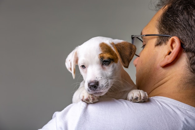 Horizontal portrait of handsome cheerful man holds jack russell terrirer on his shoulder, has glad expression