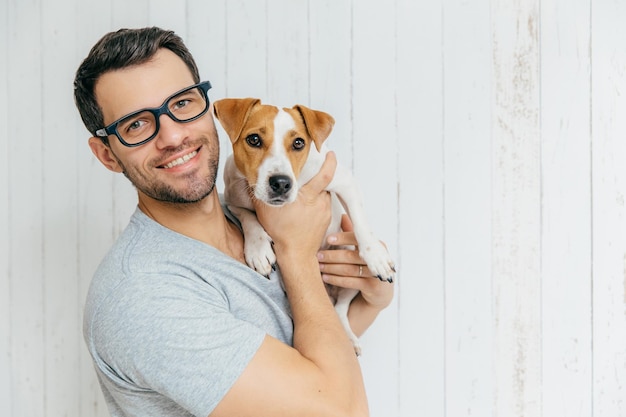 Il ritratto orizzontale dell'uomo allegro bello indossa gli occhiali tiene il terrier di jack russell ha l'espressione felice pone contro la parete di legno bianca con lo spazio vuoto della copia animali e amicizia