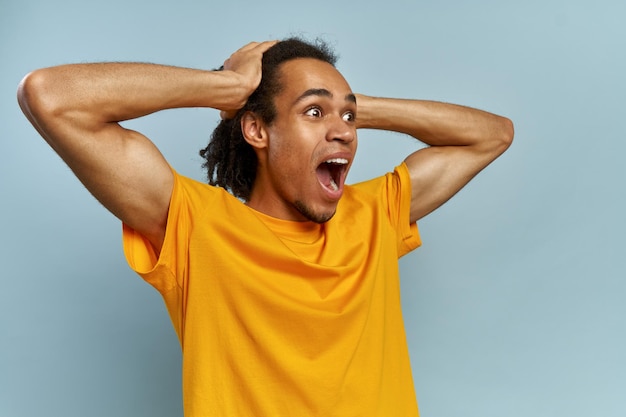 Horizontal portrait of man with dark skin and Afro hairstyle screams in despair, opens mouth widely, being in panic. Frustrated mixed race man poses against blue studio background with copy space.