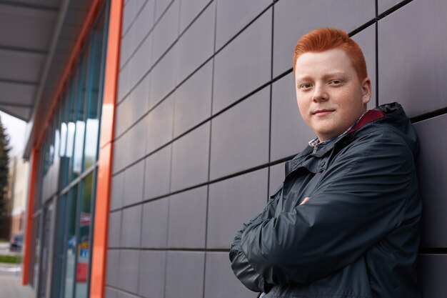 A horizontal portrait of redhead guy with freckles wearing black jacket posing over black wall