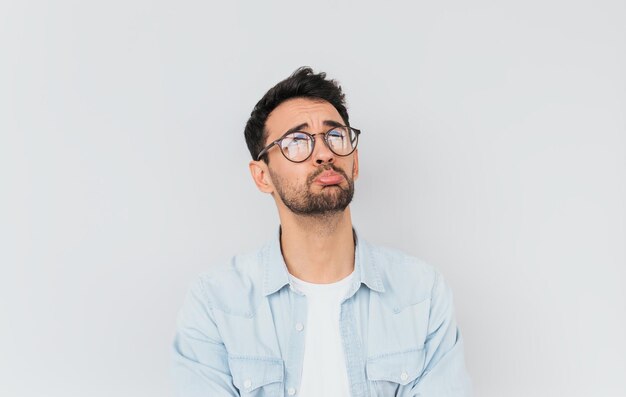 Horizontal portrait of stressed frustrated man curves lower lips isolated over white studio background Bearded male having sad expression wearing blue shirt on white Tshirt People emotions concept