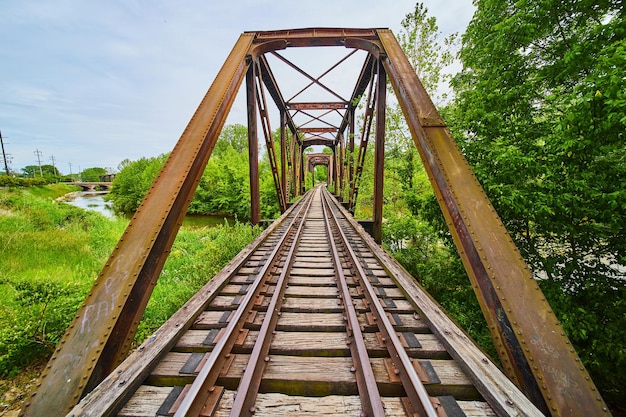 Horizontal rusty iron railroad bridge with train tracks leading into forested area over river