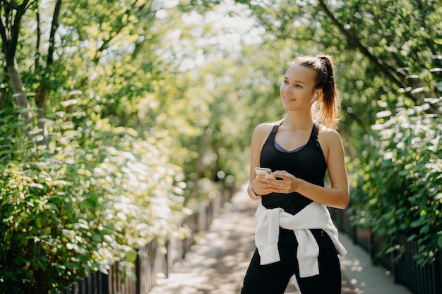 Horizontal shot of active sporty woman uses smartphone for\
checking results after jogging dressed in active wear enjoys warm\
sunny day listens music via earphones healthy lifestyle\
concept