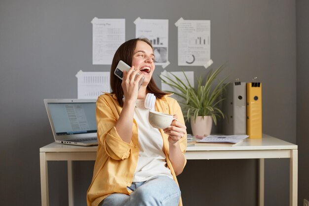 Горизонтальный снимок очаровательной красивой радостной женщины с каштановыми волосами в желтой рубашке, позирующей в офисе и разговаривающей по телефону во время перерыва, пьющей кофе или чай