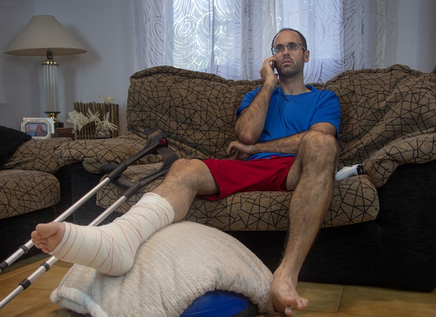 Horizontal shot of an adult man with his leg in a cast and\
bandaged on a stool with crutches at his side talking on the mobil.\
concept of rehabilitation after serious physical accident\
injury