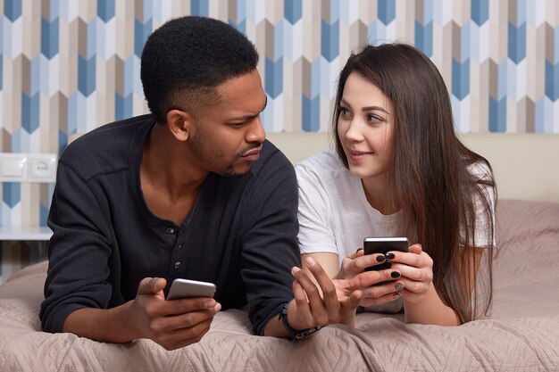 Horizontal shot of African American guy, his girlfriend, being addicted to modern technologies, lie in bed on stomach, talk about something