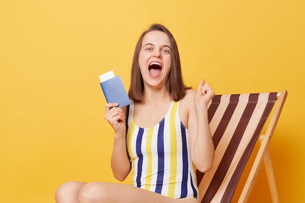 Horizontal shot of amazed excited overjoyed young woman wearing colorful swimsuit holding tickets and passport in hands yelling with happiness sitting on wooden chair isolated on yellow background