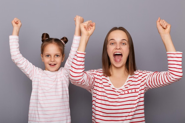 Горизонтальный снимок изумленной счастливой веселой женщины и маленькой девочки с двумя пучками волос, стоящих с поднятыми сжатыми кулаками, кричащими ура, позирующими изолированными на сером фоне