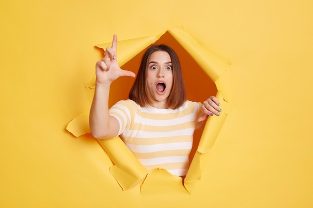 Horizontal shot of astonished surprised woman wearing striped t shirt posing standing through yellow paper torn hole looking at camera with widely opened mouth