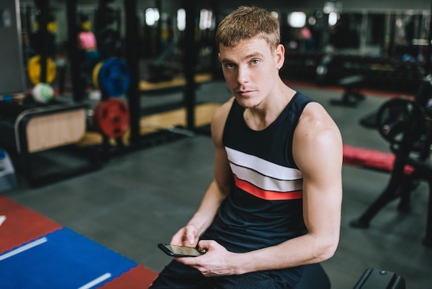 Horizontal shot of athletic man in the gym using smart phone for sport application doing fitness exercise sporty male sitting on a brench in the fitness club with copy space for text or advertising