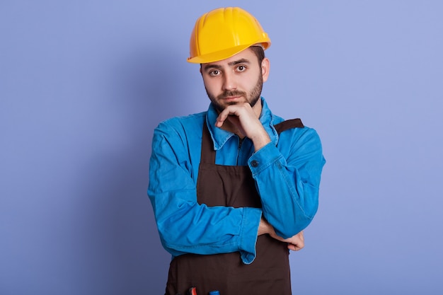 Photo horizontal shot of attentive serious young handyman having pensive facial expression, , wearing apron, helmet and overalls, having thoughts about project.