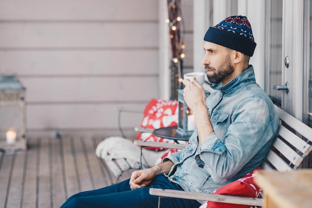 Horizontal shot of attractive bearded male wears hat and denim jacket drinks hot tea sits at bench on balcony has thoughtful expression thinks about something important in life Leisure time