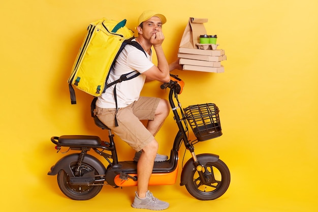 horizontal shot of attractive bored man with backpack wearing white t shirt riding electric bike posing isolated over yellow background holding food orders in hands waiting client for handing order