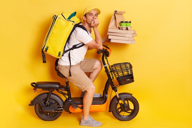 Horizontal shot of attractive bored man with backpack wearing white t shirt riding electric bike posing isolated over yellow background holding food orders in hands waiting client for handing order