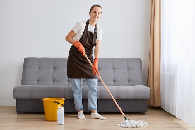 Horizontal shot of attractive Caucasian woman wearing casual clothing and brown apron washing floor holding mop and looking at camera cleaning living room