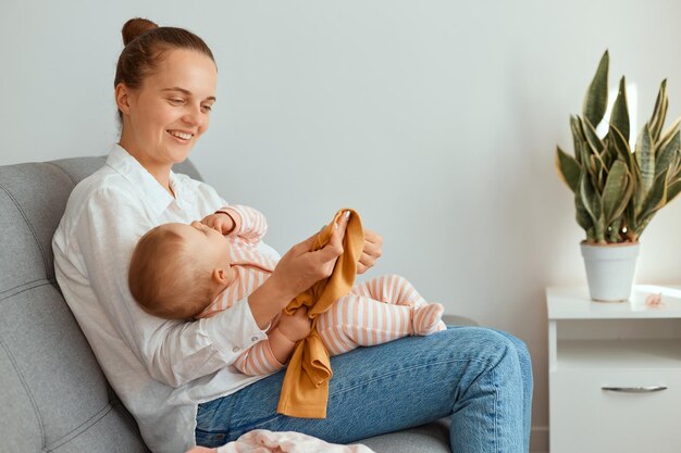 Horizontal shot of attractive young adult mother sitting on sofa with her toddler daughter, expressing happiness, smiling happily, motherhood and childhood.