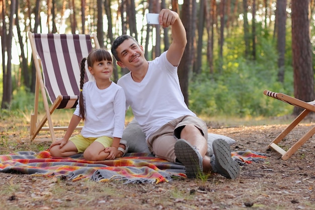 Horizontal shot of attractive young father in white t shirt sitting on blanket with little girl his daughter and taking selfie from their vacation smiling to cell phone camera enjoying rest