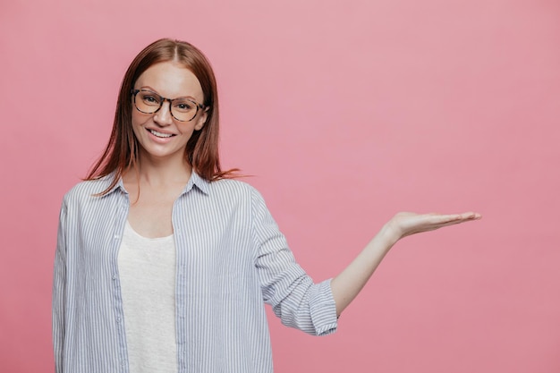 Horizontal shot of attractive young lady holds free space, dressed in formal clothes, smiles gently, poses over pink background, asdvertises new item in shop, has glad facial expression. Advertisement