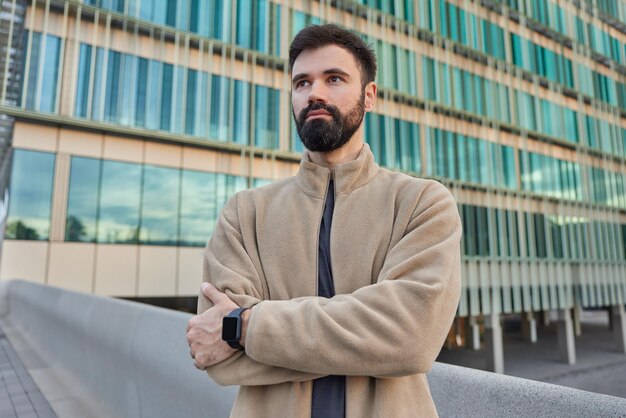 Horizontal shot of bearded man keeps arms crossed looks thoughtfully into distance being deep in thoughts poses against modern glass building in urban setting waits for friend has serious expression