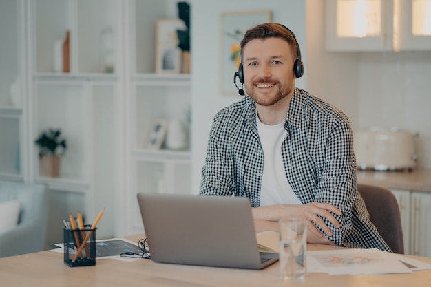 Horizontal shot of bearded man sits at desktop wears headset sits at desktop uses headphones studies or works from home prepared for video conference studies languages online poses at cozy interior