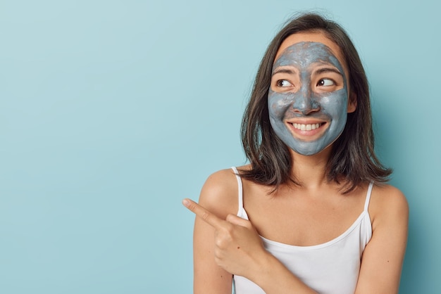 Horizontal shot of beautiful brunette asian woman with dark\
hair applies mud facial mask smiles broadly dressed in casual white\
t shirt points away on blank space for your advertising\
content