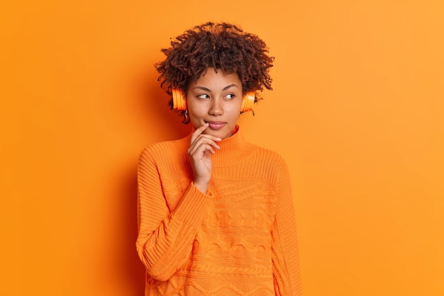 Horizontal shot of beautiful thoughtful woman with curly hair\
keeps hand near lips looks confusingly aside dressed in jumper and\
headphones isolated over vivid orange background let me think a\
bit