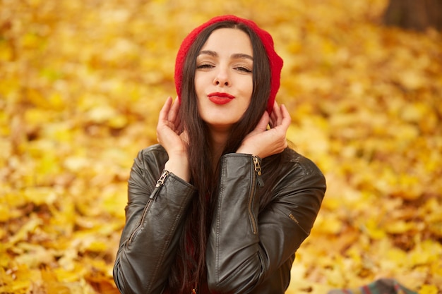 Horizontal shot of beautiful woman showing kiss gesture and smiling, posing outdoor on autumn park