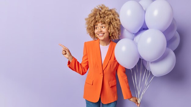 Horizontal shot of beautiful woman with curly hair dressed in
orange jacket and jeans points away on blank space invites you to
party holds bunch of helium balloons isolated over purple
background