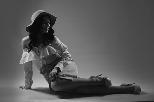 Horizontal shot black and white photo of stylish young woman in hat shirt and skirt sits on floor