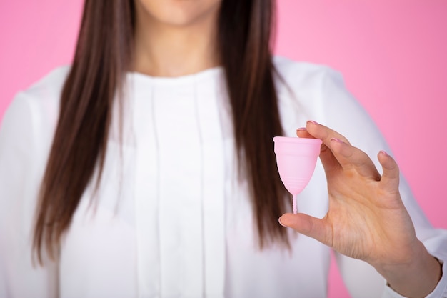 Photo horizontal shot of brunette female holding in hand plastic menstrual cup isolated over pink