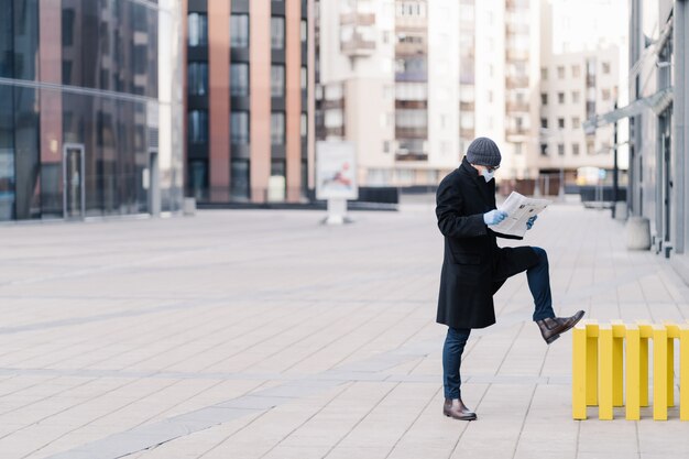 Photo horizontal shot of businessman stands outdoor near office building, reads newspaper, wears medical mask and gloves during quarantine, prevents coronavirus. pandemic situation in whole world.