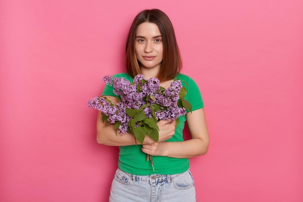 Horizontal shot of calm relaxed cute woman looks with pleasure at camera wears green t shirt holding lilac flowers posing against pink background charming female with bouquet