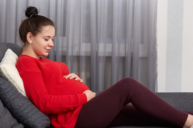 Horizontal shot of calm relaxed pregnant female wears red sweater and leggings