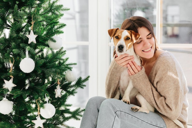 Horizontal shot of caring smiling woman hugs puppy with love enjoys cozy atmopshere and lovely moment smiles pleasantly decorate New Year tree together celebrate favourite holiday at home