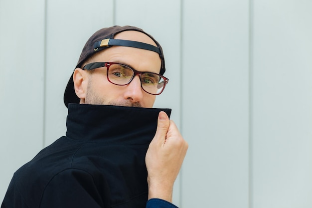 Horizontal shot of Caucasian attractive male hides behind collar of shirt wears spectacles cap has serious expression wears over white wall Serious male adult stands indoor alone Lifestyle