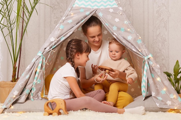 Horizontal shot of Caucasian mother with two daughters sitting together in teepee tent at home playing with wooden eco toy expressing positive emotions and happiness