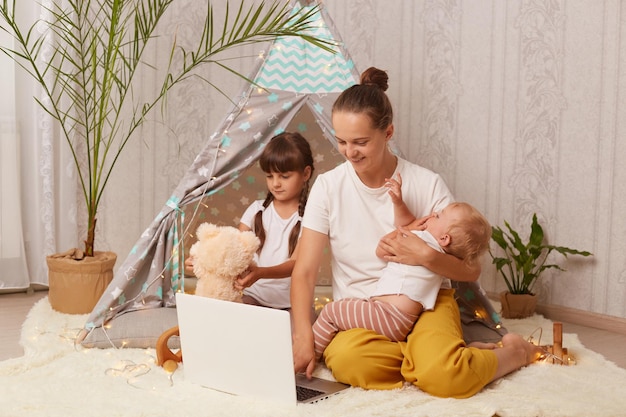 Photo horizontal shot of charming family mother with her little daughters sitting on floor near peetee tent and using laptop for watching cartoons happy childhood and motherhood