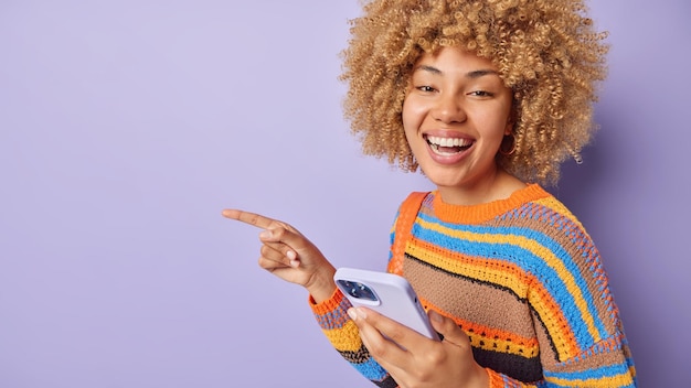 Horizontal shot of cheeful woman with curly hair points away on\
empty space shows advertisement uses mobile phone for sending sms\
dressed casually isolated over purple background look here