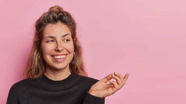 Photo horizontal shot of cheerful caucasian woman with beaming pleasant smile on face keeps hand raised concentrated aside dressed casually stands against pink background copy space for your promotion