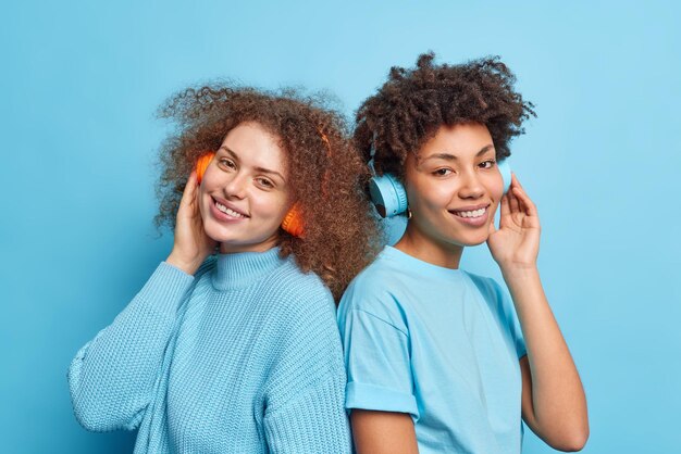 Horizontal shot of cheerful curly haired best friends stand\
back to each other enjoys listening favorite music via stereo\
headphones smile gently dressed casually isolated over blue\
background