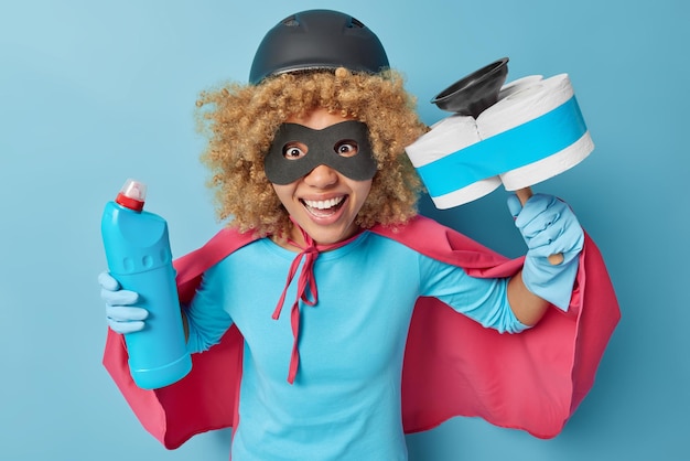 Horizontal shot of cheerful curly woman dressed like superhero\
has happy expression holds cleaning detergent and plunger going to\
wash house isolated over blue background housekeeping concept