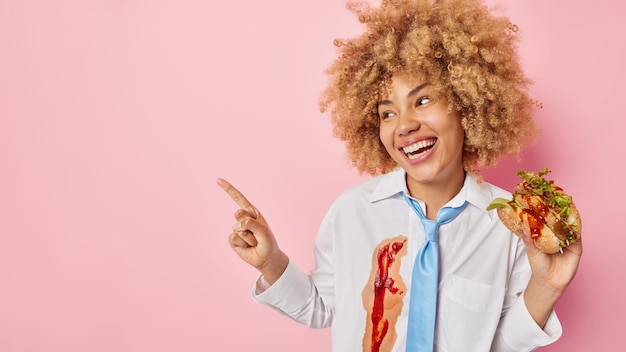 Horizontal shot of cheerful curly woman eats burger has unhealthy snack wears clothes dirty with ketchup points index finger left on empty space demonstrates something against pink background