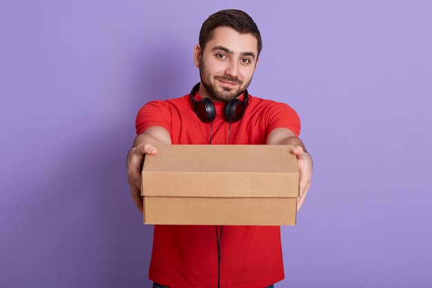 Horizontal shot of cheerful delivery man with beard. happy young courier holding cardboard box and smiling while standing against lilac wall, having headphones around neck