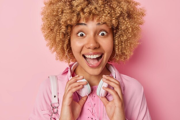 Horizontal shot of cheerful excited young woman with curly hair\
keeps hands on stereo headphones keeps mouth opened cannot believe\
in amazing news wears jacket isolated over pink background