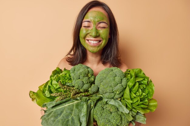 Horizontal shot of cheerful woman applies natural homemade mask\
on face for skin treamtent closes eyes and smiles toothily carries\
fresh vegetables for making vegetarian salad poses indoors