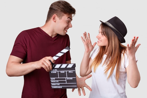 Horizontal shot of cheerful woman and man look positively at each other, gesture actively, have hesitant expressions, holds clapper board