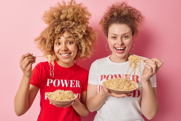 Horizontal shot of cheerful women enjoy eating delicious\
noodles for lunch stand next to each other dressed in casual t\
shirts isolated over pink background people friendship and\
nutrition concept