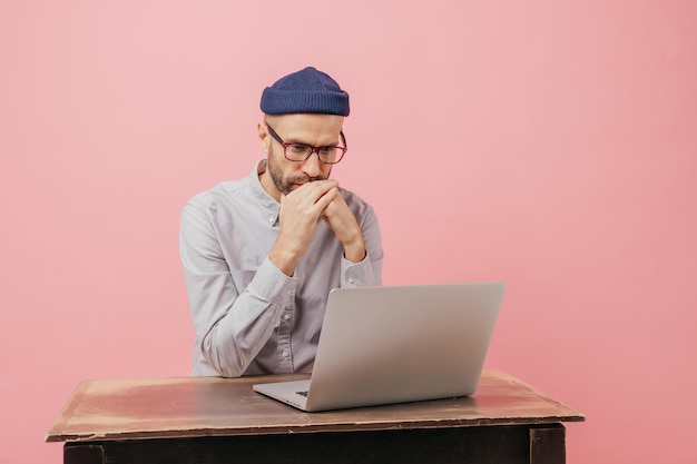 Horizontal shot of concentrated male worker reads statistics\
attentively focused in laptop computer learns something online\
wears spectacles hat and white shirt isolated over pink wall