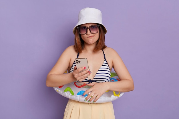 Horizontal shot of confused pensive woman with rubber ring wearing panama and swimming suit holding smart phone thinking about answer on a question posing isolated over purple background