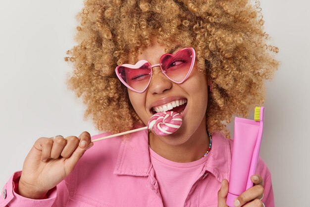 Horizontal shot of curly haired european woman bites caramel\
candy on stick winks eye holds toothbrush and toothpaste eats\
harmful food for teeth wears heart shaped sunglasses and pink\
jacket